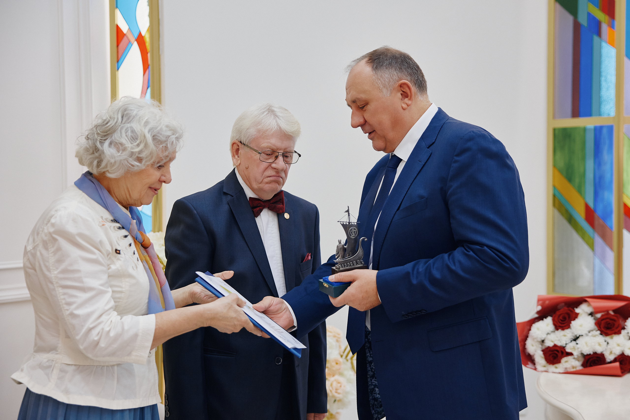 Женщина передаёт мужчине в синем костюме папку с дипломом, он держит статуэтку в виде корабля. Рядом стоит мужчина в тёмном костюме с бабочкой. За ними — светлые шторы и витражная полоса. Атмосфера торжественная, церемониальная.