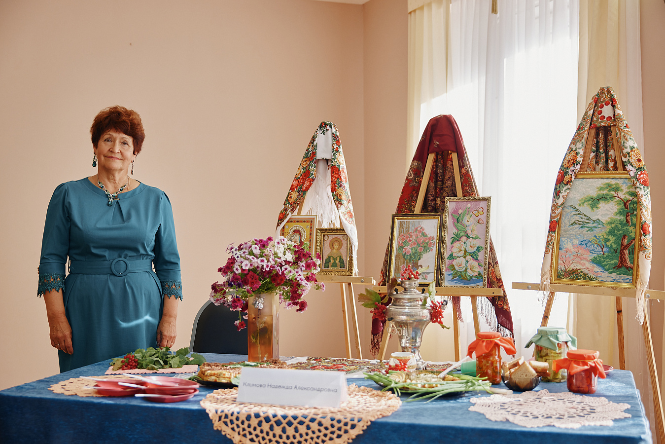 Женщина в бирюзовом платье стоит у стола с синей скатертью. На столе — вышитые картины, иконы, самовар, цветы, угощения и рушники. Всё оформлено в народном стиле. Атмосфера тёплая, домашняя, праздничная.