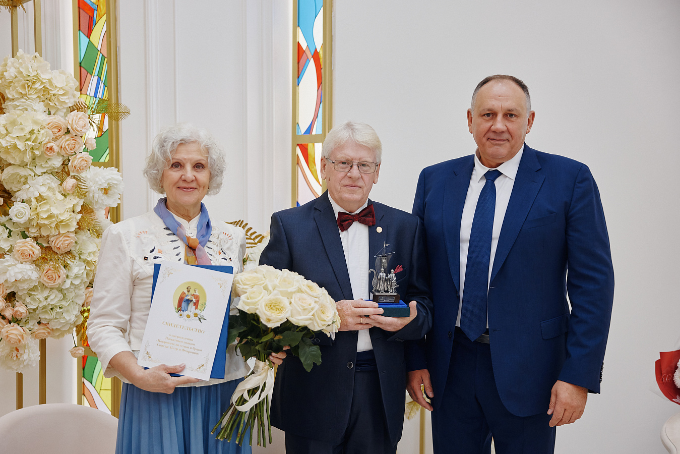 Трое людей на сцене: женщина с дипломом и букетом белых роз, мужчина с бабочкой и статуэткой в руках, и мужчина в синем костюме. За ними — светлые шторы и цветная витражная полоса, слева — композиция из белых цветов. Атмосфера торжественная, праздничная.